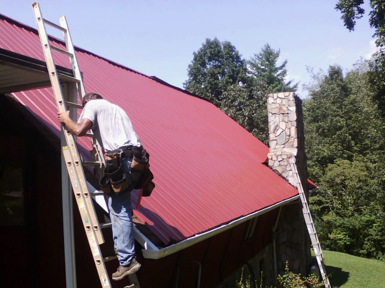 Roofer on a ladder beside a red metal roof and stone chimney — Soffit & Fascia Repair by Roofing Brothers in Hometown